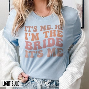 May include: Light blue t-shirt with the text "It's me, hi I'm the bride it's me" in a retro font with orange and pink color gradient.