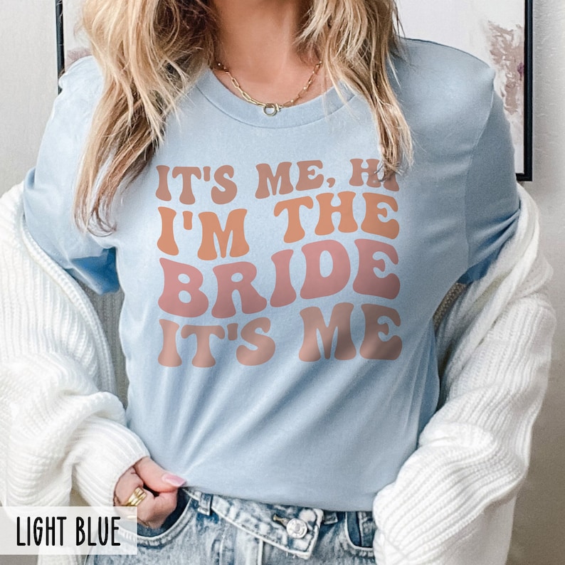 May include: Light blue t-shirt with the text "It's me, hi I'm the bride it's me" in a retro font with orange and pink color gradient.