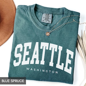 May include: Blue spruce t-shirt with the word "SEATTLE" in large white block letters, with "WASHINGTON" underneath. A gold necklace with a pendant rests on the shirt. The shirt is folded and has a "Comfort Colors" tag.