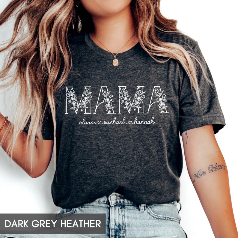 May include: Dark grey heather t-shirt with the word "MAMA" in white, floral-filled letters. Below the word "MAMA" are the names "olivia", "michael", and "hannah" with hearts. The shirt is a v-neck style.