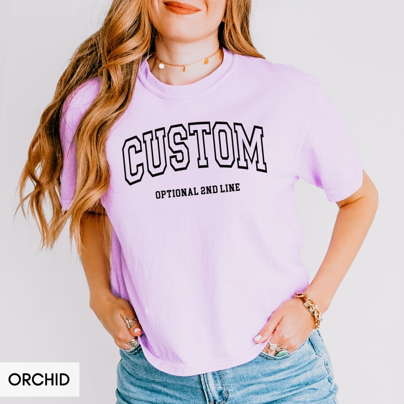 May include: A light purple t-shirt with the word "CUSTOM" in a black varsity-style font. The text "OPTIONAL 2ND LINE" is printed below the word "CUSTOM".