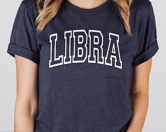 May include: A heather navy blue short-sleeve t-shirt featuring the word LIBRA printed in a large, white, arched collegiate-style font across the chest. The garment has a crew neckline and cuffed sleeves, showcasing a textured fabric finish.