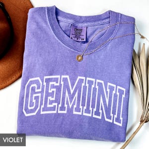 May include: A violet Comfort Colors t-shirt with the word "GEMINI" in white block letters. A gold necklace with a pendant rests on the shirt. A brown hat and dried plant stems are in the background.