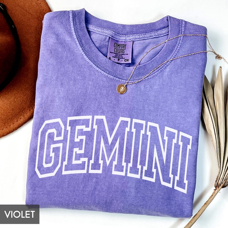 May include: A violet Comfort Colors t-shirt with the word "GEMINI" in white block letters. A gold necklace with a pendant rests on the shirt. A brown hat and dried plant stems are in the background.