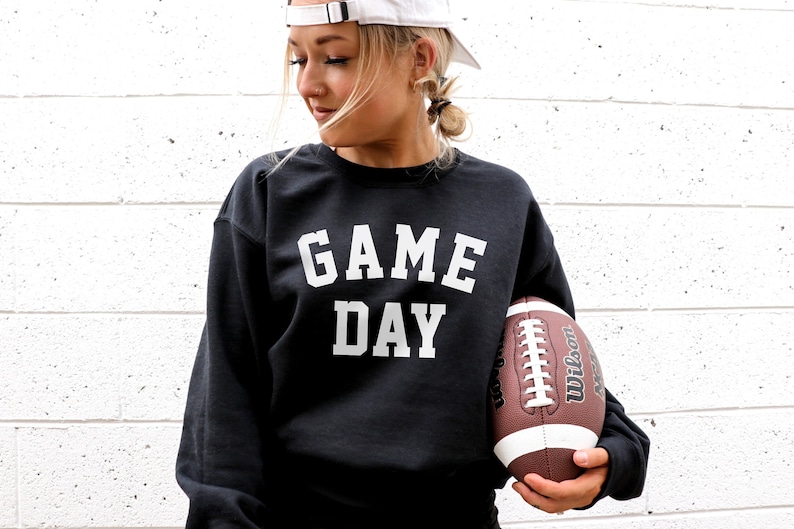 May include: A black sweatshirt with the text "GAME DAY" in white lettering. A person is holding a brown and white football.
