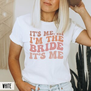 May include: White t-shirt with the text "It's me, I'm the bride, it's me" in a retro font with pink, orange, and red gradient.