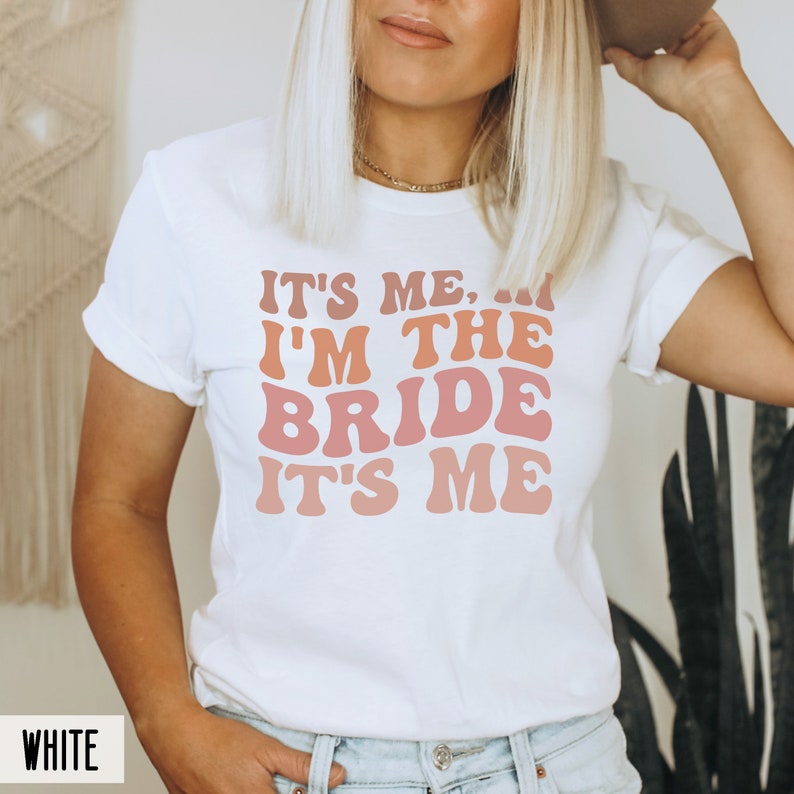 May include: White t-shirt with the text "It's me, I'm the bride, it's me" in a retro font with pink, orange, and red gradient.