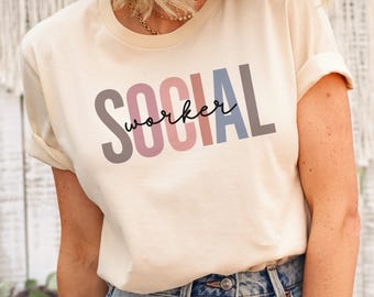 May include: A soft cream-colored short-sleeve t-shirt featuring the word SOCIAL in large, multi-colored block letters and the word worker written in a black cursive script across the center of the chest. The shirt has a crew neckline.