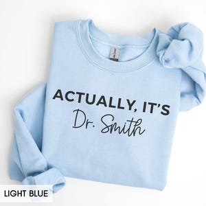 May include: Light blue sweatshirt with the text "ACTUALLY, IT'S Dr. Smith" in black. The sweatshirt is a crew neck style and the words "LIGHT BLUE" are at the bottom of the image.