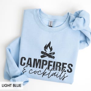 May include: Light blue sweatshirt with the words "Campfires & cocktails" in black. The design includes a campfire graphic. The sweatshirt is a crew neck style.