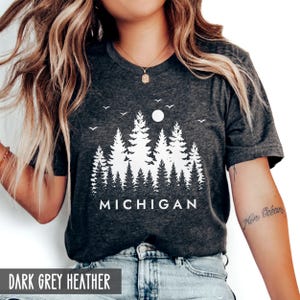 May include: A dark grey heather t-shirt featuring a white graphic of a forest with pine trees, a sun, and birds. The word MICHIGAN is printed in white capital letters below the trees. The shirt has short sleeves and a crew neckline.