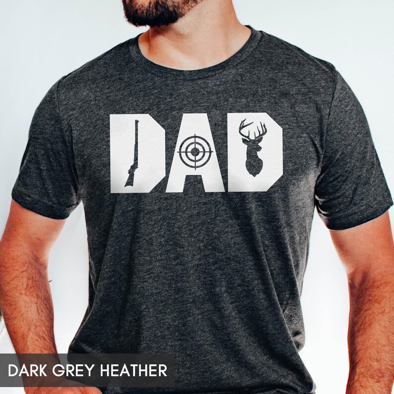 May include: Dark grey heather t-shirt with the word "DAD" in white block letters. The "D" features a rifle, the "A" a target, and the second "D" a deer silhouette. The text "DARK GREY HEATHER" is at the bottom.