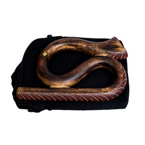 May include: A handcrafted wooden didgeridoo with a curved design, resting in a black carrying case. The didgeridoo features a dark brown finish with intricate dot patterns in red, yellow, and white. The case has a zipper closure.