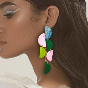 Könnte beinhalten: Ein Paar lange, farbenfrohe Ohrringe mit geometrischem Design. Die Ohrringe bestehen aus grünen, rosa und blauen Halbkreisen und sind durch eine goldene Kette verbunden.