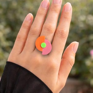 May include: A colorful ring with a geometric design. The ring features a half-circle in orange, a half-circle in pink, and a small green circle. The ring is worn on a finger.