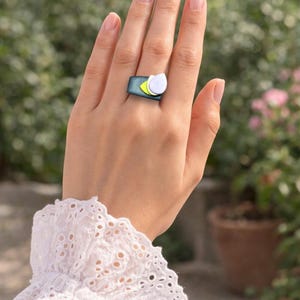 May include: A unique ring featuring a rectangular teal band with a yellow accent and a white, petal-shaped design. The ring is worn on a hand with manicured nails, set against a blurred green and pink background.