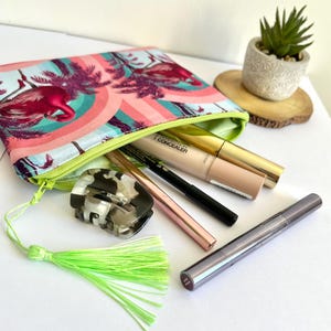 Può includere: Una pochette con cerniera rosa e verde con stampa di fenicottero e palme. La pochette è aperta e contiene pennelli per il trucco, un correttore e una forcina per capelli.