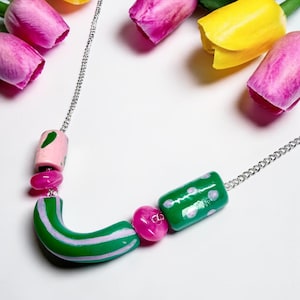 May include: A silver chain necklace featuring a variety of colorful beads, including a pink cylindrical bead, a green cylindrical bead with white dots, two small translucent pink beads, and a curved green bead with light pink stripes.
