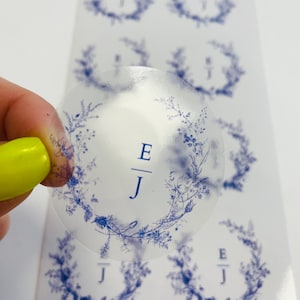 Könnte beinhalten: Transparente runde Aufkleber mit einem blauen floralen Kranzdesign und den Initialen "E/J" in der Mitte.