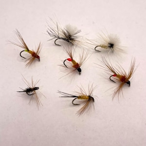 Può includere: Una collezione di sette mosche da pesca artificiali. Le mosche presentano vari colori, tra cui marrone, nero, bianco e giallo, con accenti rossi. Ogni mosca ha un piccolo amo e ali piumate, progettate per imitare gli insetti.