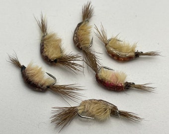May include: Six brown and white fly fishing lures with feathered tails. The lures are tied on hooks and have a realistic shrimp design.