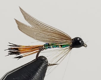May include: A close-up of a handmade fishing fly. The fly features a black head, iridescent green and blue body, and tan wings. The tail is a mix of brown and orange fibers. The fly is attached to a silver hook.