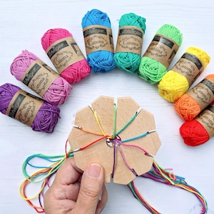 May include: A collection of colorful yarn skeins in shades of purple, pink, blue, green, yellow, orange, and red, arranged around a wooden bracelet-making tool. Each yarn ball has a brown label with the text "Scheepjes Catona".