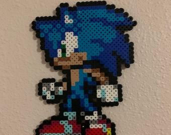 May include: A blue and white pixel art character of Sonic the Hedgehog, made with perler beads. The character is standing with his arms at his sides and his legs slightly apart. He is wearing red shoes with white laces.