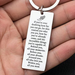 May include: A silver-colored rectangular keychain with a circular keyring. The keychain has engraved text and a winged emblem. The text reads: "If you're ever doubting how far you can go...all you need."