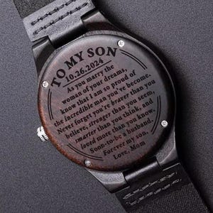 May include: A wooden wristwatch with a black leather band. The watch face is engraved with a heartfelt message to a son, including the date 10.26.2024 and the words "To My Son" at the top. The message is from "Mom."