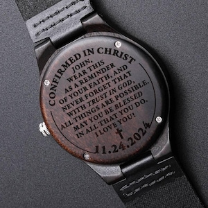 May include: A wooden wristwatch with a dark brown face and black leather strap. The watch face is engraved with the words "CONFIRMED IN CHRIST" and a religious message. The date "11.24.2024" is also engraved on the watch face.