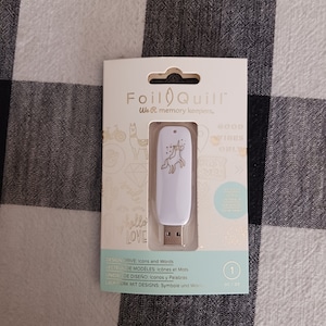 Puede incluir: Unidad USB blanca con un diseño de unicornio dorado. La unidad está empaquetada en una caja blanca con el texto "Foil Quill We R memory keepers." y "Design Drive: Iconos y Palabras".