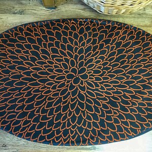 Puede incluir: Una alfombra redonda de color negro y naranja con un patrón circular de pétalos naranjas sobre un fondo negro.