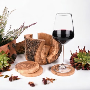 Könnte beinhalten: Ein Weinglas mit Rotwein steht auf einem Untersetzer aus Holz. Ein hölzerner Weinglashalter und mehrere Untersetzer aus Holz sind ebenfalls vorhanden. Die Artikel sind mit dekorativen Pflanzen und Blättern angeordnet.