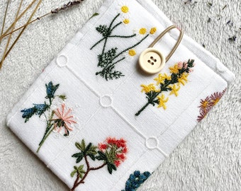 Puede incluir: Funda de teléfono de tela blanca con flores bordadas en tonos de rosa, amarillo, verde y azul. La funda tiene un cierre de botón blanco.