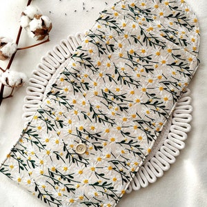 May include: A white fabric pouch with a floral pattern of white daisies and green stems. The pouch has a button closure and a loop handle.