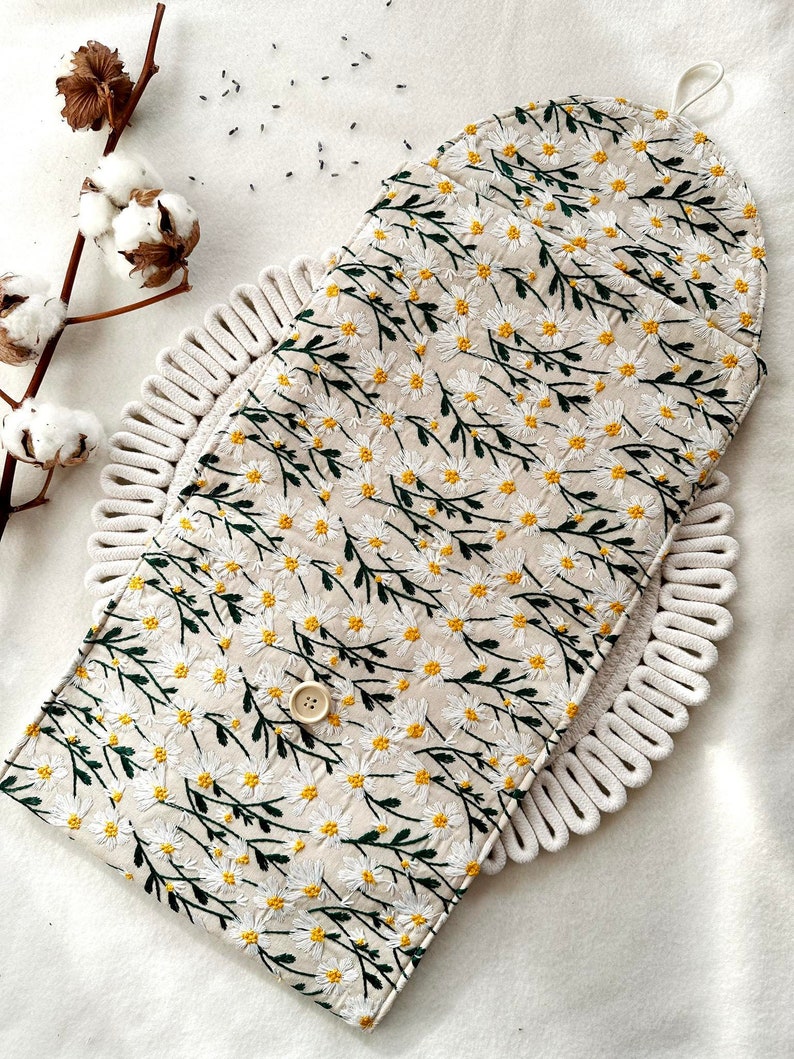 May include: A white fabric pouch with a floral pattern of white daisies and green stems. The pouch has a button closure and a loop handle.