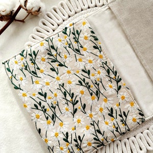 May include: A white linen clutch with a floral embroidery design. The embroidery features small white daisies with yellow centers and green stems. The clutch has a rope trim around the edges.