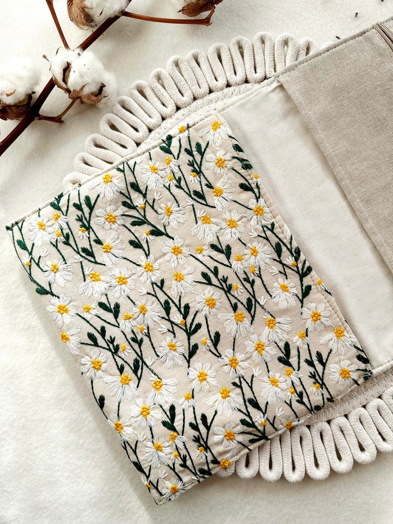 May include: A white linen clutch with a floral embroidery design. The embroidery features small white daisies with yellow centers and green stems. The clutch has a rope trim around the edges.