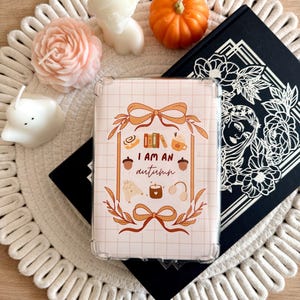 May include: A clear protective case for an electronic device featuring a grid pattern and a central graphic with the text I AM AN autumn surrounded by illustrations of a cinnamon roll, books, a mug, acorns, a sweater, a candle, and headphones.