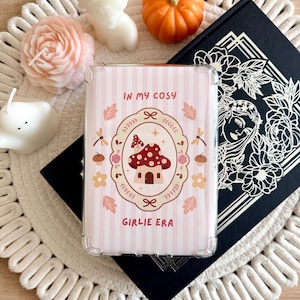 May include: A rectangular clear protective case featuring a light pink and white striped background. The center displays an oval frame containing a red mushroom house, surrounded by small flowers, leaves, and dragonflies. Text reads IN MY COSY GIRLIE ERA.