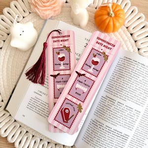 May include: Two rectangular paper bookmarks with a pink and white striped background. Each features three stacked illustrations: a glass of bloody mary, vampire fangs, and a coffin with the text Summerween Date Night, A few bloody marys, A Fang-tastic book, and Eternal rest.