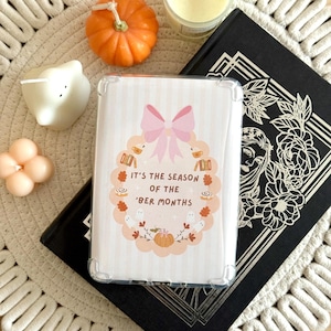 Op de afbeelding: Een doorzichtige telefoonhoes met een herfstontwerp, met een roze strik, pompoenen, geesten en de tekst "IT'S THE SEASON OF THE 'BER MONTHS". De hoes ligt op een zwart boek, omringd door een kleine pompoen, kaarsen en een geweven placemat.