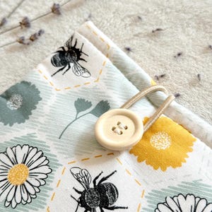 May include: A white fabric pouch with a bee and daisy pattern. The pouch has a wooden button closure and a beige drawstring.