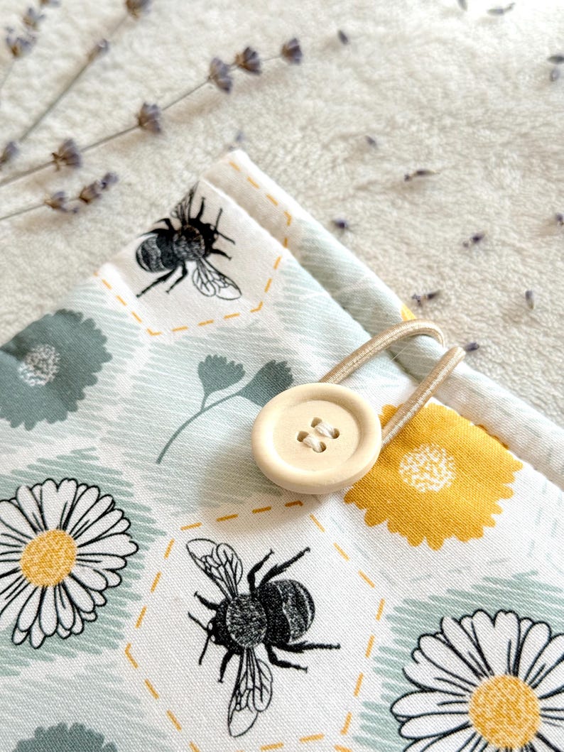 May include: A white fabric pouch with a bee and daisy pattern. The pouch has a wooden button closure and a beige drawstring.