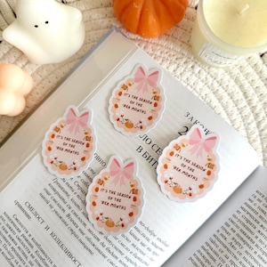 May include: Four circular stickers with a pink bow and the text "IT'S THE SEASON OF THE 'BER MONTHS" are displayed on an open book. The stickers are surrounded by pumpkins, leaves, and other fall-themed elements. A small pumpkin, a candle, and a ghost-shaped candle are also in the image.