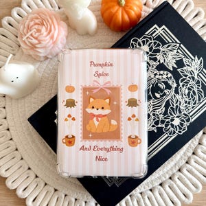 May include: A clear protective case for a device featuring a light pink and white striped background with a central illustration of a fox, pumpkins, tree stumps, and mugs. The text reads Pumpkin Spice And Everything Nice in brown font.