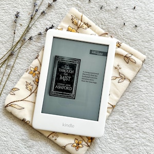 May include: A white Kindle e-reader with a gray screen displaying the cover of the book "Through the Mist" by Lindsay Jayne Ashford. The book cover features a dark gray background with a white house and the title in white text. The Kindle is resting on a white fabric with a floral pattern.