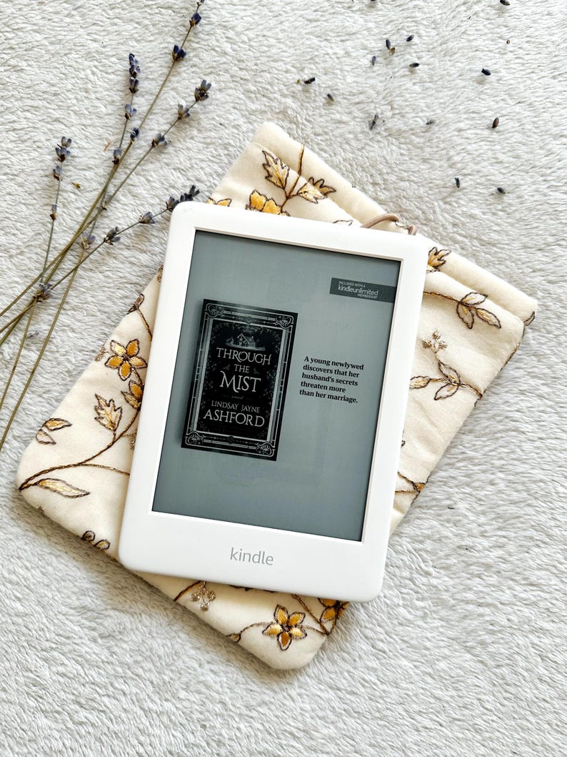 May include: A white Kindle e-reader with a gray screen displaying the cover of the book "Through the Mist" by Lindsay Jayne Ashford. The book cover features a dark gray background with a white house and the title in white text. The Kindle is resting on a white fabric with a floral pattern.