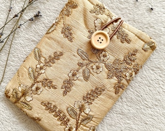 Puede incluir: Una bolsa de tela beige con un patrón floral y bordados dorados. La bolsa tiene una correa de cuero marrón y un cierre de botón de madera.
